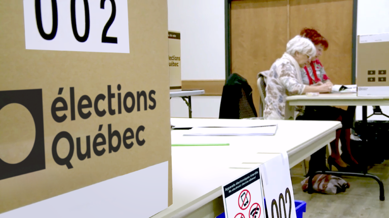 Gros plan d’une urne numérotée, avec le logo d’Élections Québec, sur une table. On voit également des membres du personnel, deux femmes, qui vérifient des documents sur une autre table à l’arrière-plan.