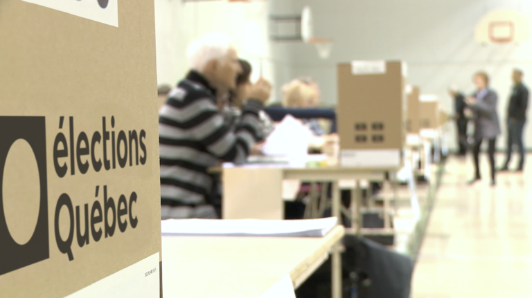 Gros plan d’une urne, avec le logo d’Élections Québec, sur une table. Le reste de l’image est flou et montre des membres du personnel et des électeurs.