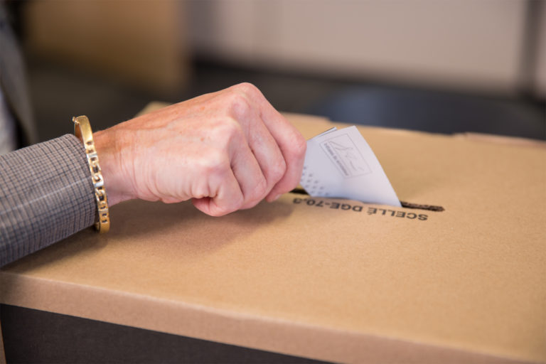 Gros plan d’une main de femme âgée qui dépose son bulletin de vote dans une urne, à gauche.