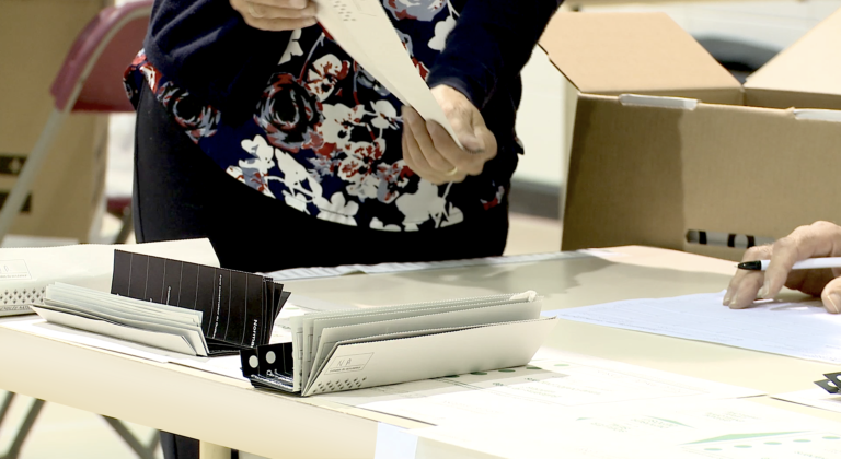 Gros plan d’une table sur laquelle se trouvent des piles de bulletins de vote dépouillés derrière laquelle un membre du personnel montre un bulletin de vote à un collègue hors champ.