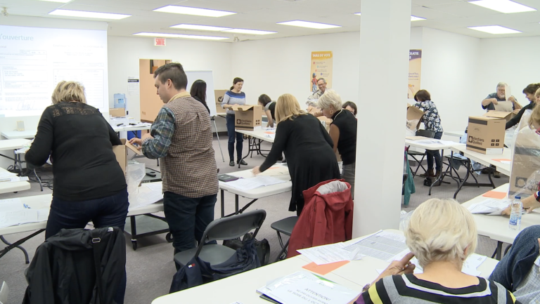 Plan large d’une salle bondée de gens de tout âge affairés autour de tables sur lesquelles se trouvent des documents et des urnes lors d’une formation pour le personnel électoral.