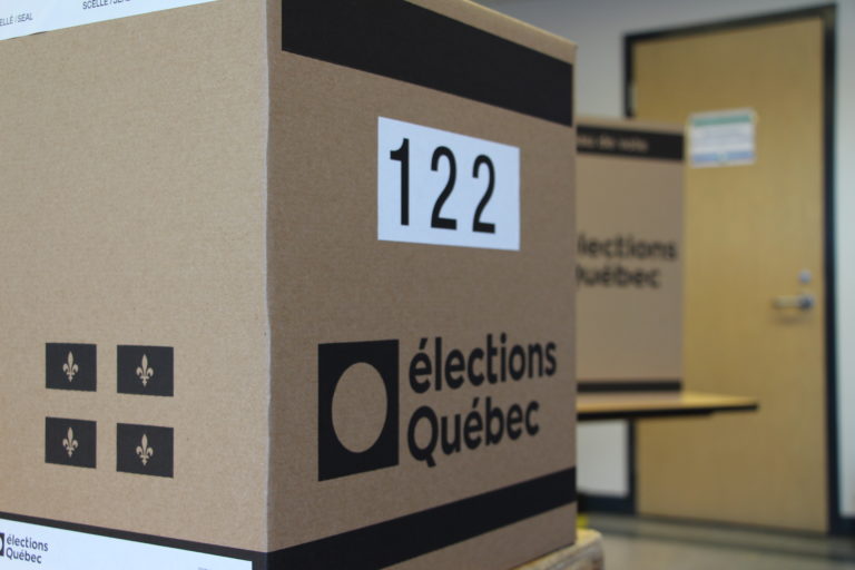 Vue de trois quarts d’une urne. On peut voir le logo d’Élections Québec et le numéro de l’urne d’un côté ainsi que le drapeau du Québec sur l’autre côté. En arrière-plan, il y a un isoloir.