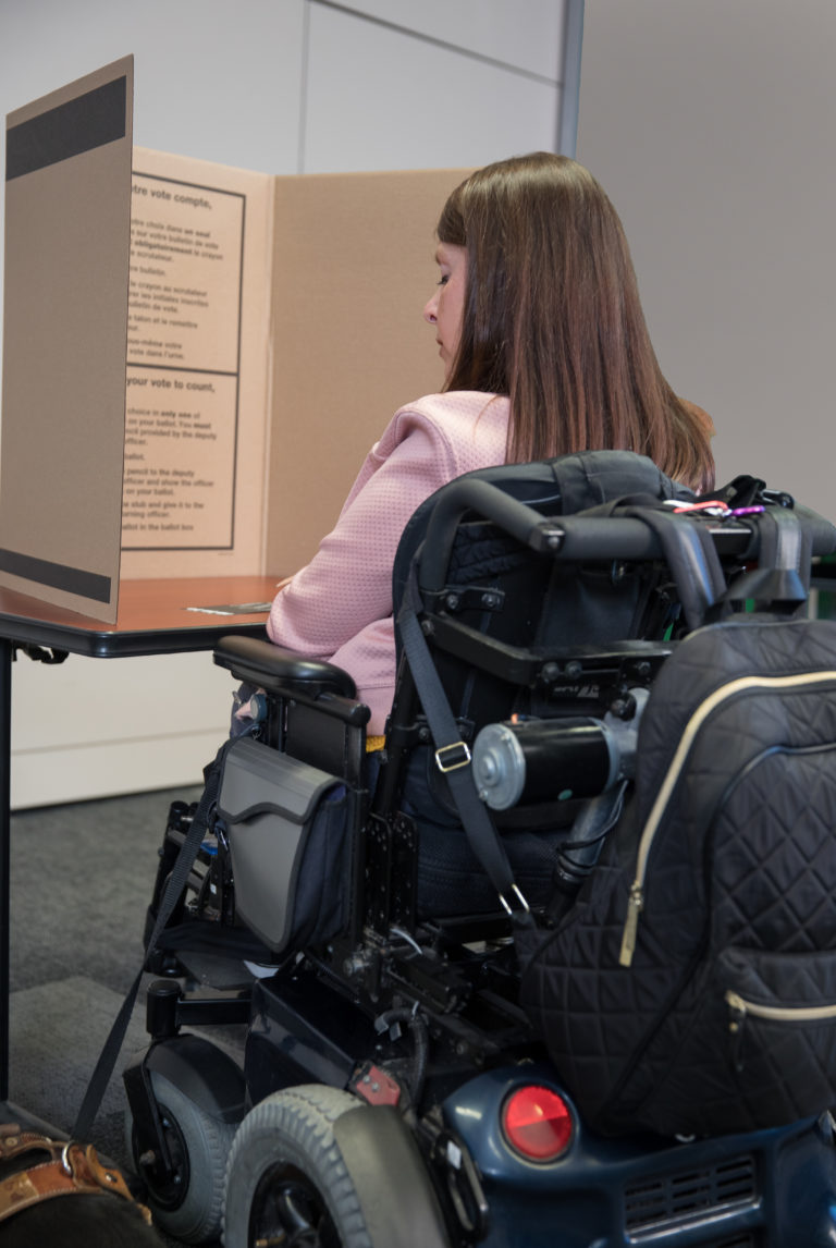 On voit une femme assise dans un fauteuil roulant électrique, de dos, avec son bulletin de vote dans un isoloir.